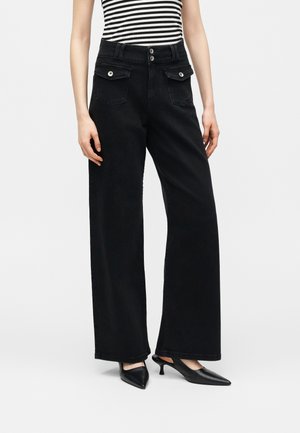 Femme portant un pantalon noir taille haute à jambes larges avec poches à rabat sur le devant et détails à boutons, associé à des escarpins noirs à bouts pointus et talons petits.