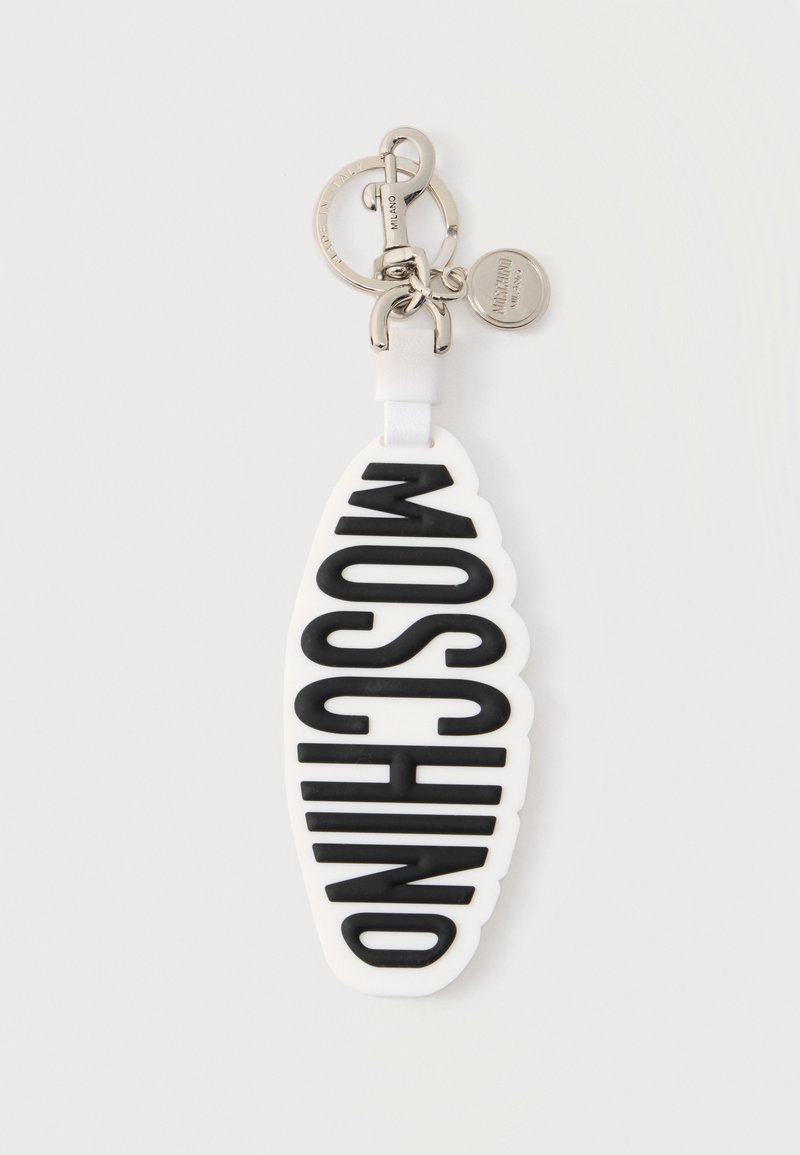 Μπρελόκ με λευκή οβάλ ετικέτα που φέρει ανάγλυφο μαύρο κείμενο "MOSCHINO", προσαρτημένο σε ασημένιο μεταλλικό δαχτυλίδι και κούμπωμα με μικρό κυκλικό γούρι.