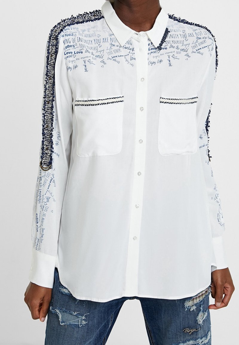 Witte button-up shirt met getextureerde blauwe borduursels en bedrukte tekst op de voorkant en mouwen. Twee voorzakken, lange mouwen.