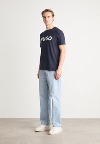 Tricou din bumbac de culoare bleumarin cu logo-ul alb "HUGO", asociat cu blugi decolorati de nuanta bleu deschis si adidași albi. Design simplu, casual, cu o croială lejeră.