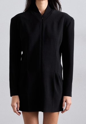 Frau trägt ein schwarzes, langärmliges, tailliertes Blazer-Kleid mit Schulterpolstern und einem Reißverschluss vorne vor einem einfarbigen hellen Hintergrund.