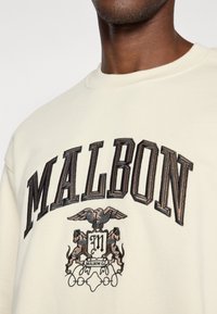 Κρεμ σακάκι με σκούρο κεντημένο κείμενο "MALBON" και έμβλημα που απεικονίζει ένα πουλί και δύο λιονταρίσιες φιγούρες. Μαλακή υφή, στρογγυλή λαιμόκ�οψη.