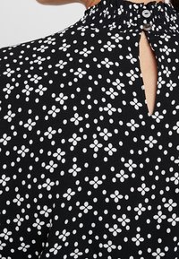 Svart tyg med vita blommönster och prickar, med en volangkrage och en liten knappstängning bak.