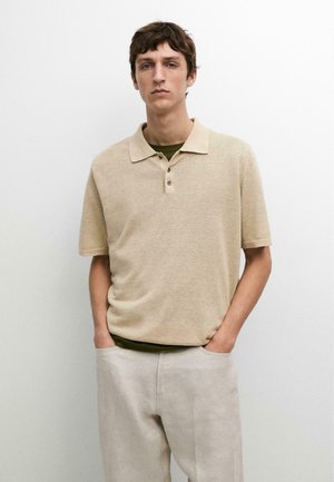 Giovane uomo che indossa una polo beige a maniche corte e pantaloni chiari, in piedi con le mani nelle tasche contro uno sfondo bianco.