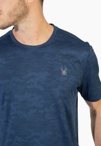 Námořnická modrá tričko s krátkým rukávem s jemným maskáčovým vzorem a malým stříbrným logem pavouka na levé straně hrudi.