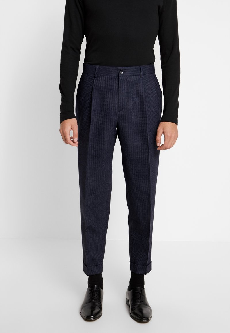Homme portant un pantalon bleu marine cintré avec revers, des chaussures habillées noires, des chaussettes noires et un haut noir à manches longues, debout sur un fond blanc.