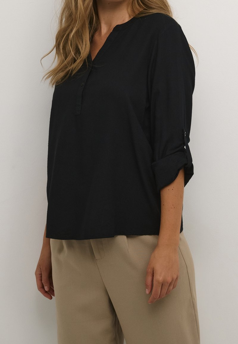 Femme portant un chemisier noir à manches longues avec manches retroussées et un pantalon beige sur un fond blanc uni.