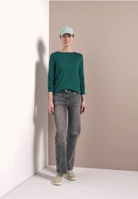 Langemouwen teal shirt, grijze jeans en een lichtgroene pet. Model staat tegen een neutrale achtergrond en toont casual kleding en schoeisel.