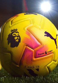 Keltainen Englannin Valioliigan jalkapallo, jossa musta leijonalogolla ja Puman tunnuksella, nurmella kirkkaan stadionvalon alla.