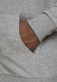 Hoodie gris en tissu doux et texturé, avec une poche kangourou et des poignets côtelés, montrant une main à l'intérieur de la poche.