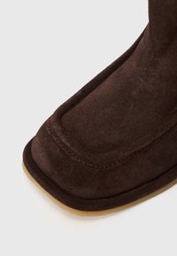 Botine din suede maro, cu vârf rotund, detalii cusute și o talpă din cauciuc de culoare bej deschis. Textură netedă cu un finisaj moale.