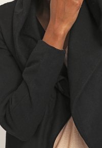 Cappotto nero, morbido e strutturato con un ampio colletto e una vestibilità comoda, che copre parzialmente una maglietta beige. Presenta un tessuto piegato sul collo.