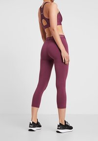 Bordowy sportowy biustonosz z wycięciem na plecach, zestawiony z bordowymi legginsami typu capri. Strój charakteryzuje się dopasowanym krojem i trwałą strukturą tkaniny. Czarne buty sportowe dopełniają wygląd.