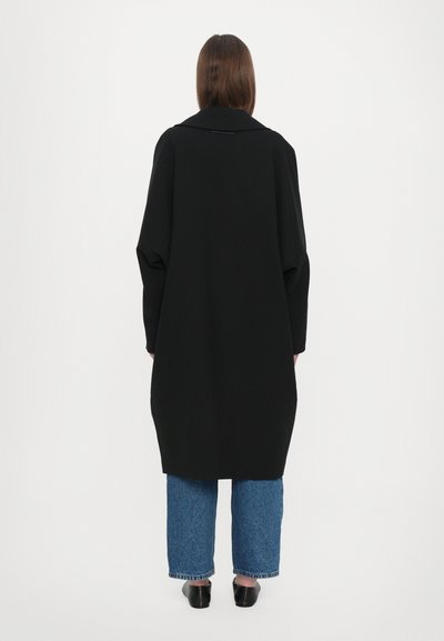 Svart oversized kappa med bred krage, långa ärmar och ett slätt, texturerat tyg; matchad med lösa blå jeans och svarta skor.