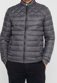 Grijze doorgestikte jas met een hoge kraag, ritssluiting en zijzakken. Heeft een contrasterende halsdetail met rode en blauwe strepen.