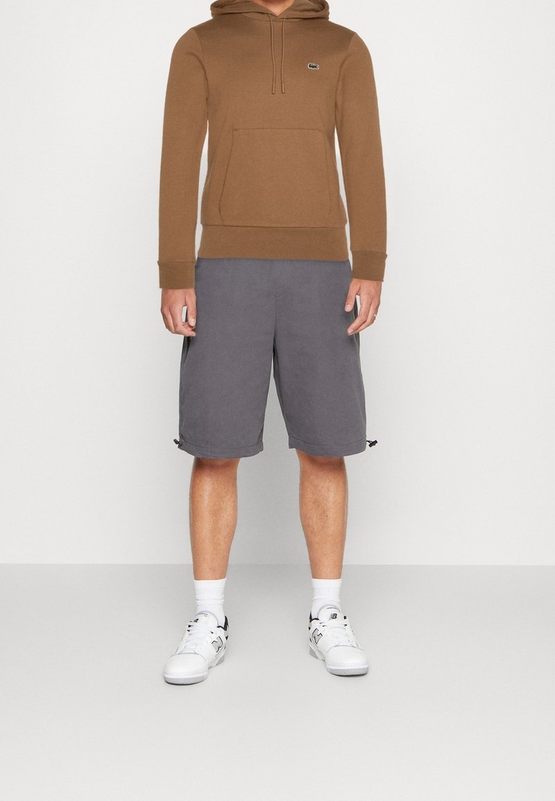 Homme portant un sweat à capuche marron, un short gris au genou, des chaussettes blanches et des baskets blanches, debout sur un fond uni.
