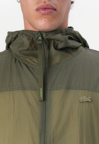 Jachetă ușoară de culoare verde măslin, cu guler înalt, fermoar frontal pe toată lungimea și material texturat. Are un logo subtil pe piept, de culoare corespunzătoare.