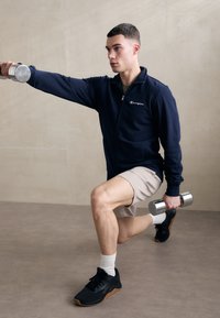 Námořnická zipová bunda, béžové kraťasy, černé sportovní boty; muž provádějící výpad s jednoručkami v každé ruce, soustředěný výraz, hladké pozadí.