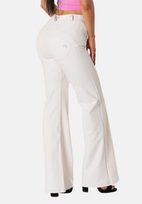 Valkoiset levenevät housut, joissa on yksityiskohtaiset ompeleet ja takataskut, joissa logo, yhdistettynä mustiin sandaaleihin. Sileä kangaspinta.