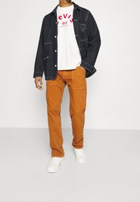 Denimjacke mit blauer Stickerei, weißes T-Shirt mit rotem Logo und orangefarbene, lässige Hose. Weiße Sneakers runden das Outfit ab.