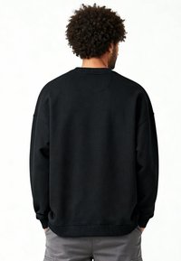 Homme aux cheveux bouclés portant un sweat-shirt noir uni et un pantalon gris, debout de dos sur un fond blanc.
