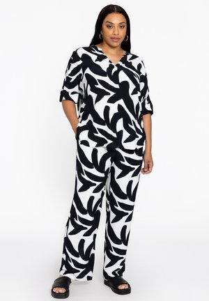 Schwarzer und weiß gemusteter Jumpsuit mit kurzen Ärmeln, V-Ausschnitt, lockerer Passform und seitlichen Taschen, kombiniert mit schwarzen Riemensandaletten.