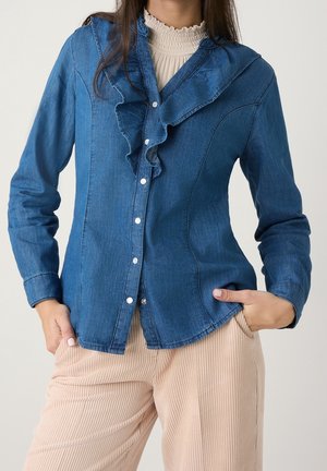 Kobieta w niebieskiej jeansowej koszuli zapinanej na guziki z marszczonym kołnierzykiem oraz beżowych spodniach sztruksowych, ręce w kieszeniach, na jednolitym tle.