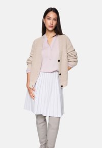 Beige gestrickte Strickjacke mit Knöpfen, blassrosa Bluse und weißem, plissiertem Rock mit vertikalen Streifen. Das Model trägt graue, figurbetonte Hosen.