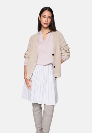 Beige gestrickte Strickjacke mit Knöpfen, blassrosa Bluse und weißem, plissiertem Rock mit vertikalen Streifen. Das Model trägt graue, figurbetonte Hosen.