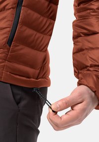 Veste matelassée couleur rouille avec un design matelassé. Présente une fermeture éclair noire et un cordon de taille ajustable, montrant une main tenant le cordon.