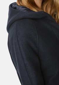Cappotto a cappuccio blu scuro con un tessuto texturizzato, cuciture visibili e una fodera interna liscia. Presenta una vestibilità comoda e hardware minimal.