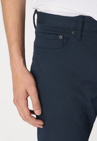 Un pantalon en coton bleu marine présente une coupe slim, des poches avant et des finitions métalliques. Le tissu a une texture lisse avec des détails de couture subtils.