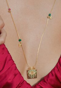 Collier en chaîne dorée avec un pendentif carré comportant un motif d'œil, orné de pierres vertes et transparentes, complété par des accents colorés.