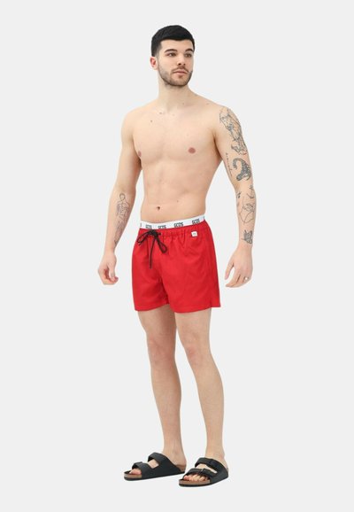 Pantaloncini da bagno rossi con vita elasticizzata e cordoncino, caratterizzati da un accento con logo, abbinati a sandali neri con suola piatta. Texture liscia.
