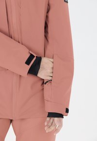 Veste imperméable rose avec fermeture éclair et poignets à Velcro, dotée d'une poche cachée et d'une doublure intérieure noire contrastante, texture lisse.