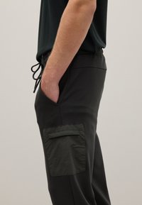 Schwarze Jogginghosen mit elastischem Bund und Kordelzug, ausgestattet mit einer Seitentasche mit einer strukturierten, dunkelgrünen Klappe. Glatte Stoffoberfläche.