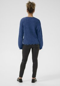Blauer Strickpullover mit weiten Ärmeln und gewelltem Saum, kombiniert mit schwarzen strukturierten Hosen und schwarzen Schuhen. Schlichtes, funktionales Design.