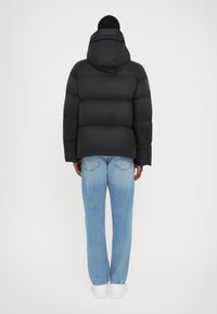 Sort pufferjakke med hætte, der har quiltet polstring og en boxy pasform, båret over lyseblå denimjeans og hvide sneakers.