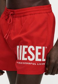Червени шорти за плуване с бяло лого Diesel и текст "Successful Living", носени от човек с тъмна кожа.