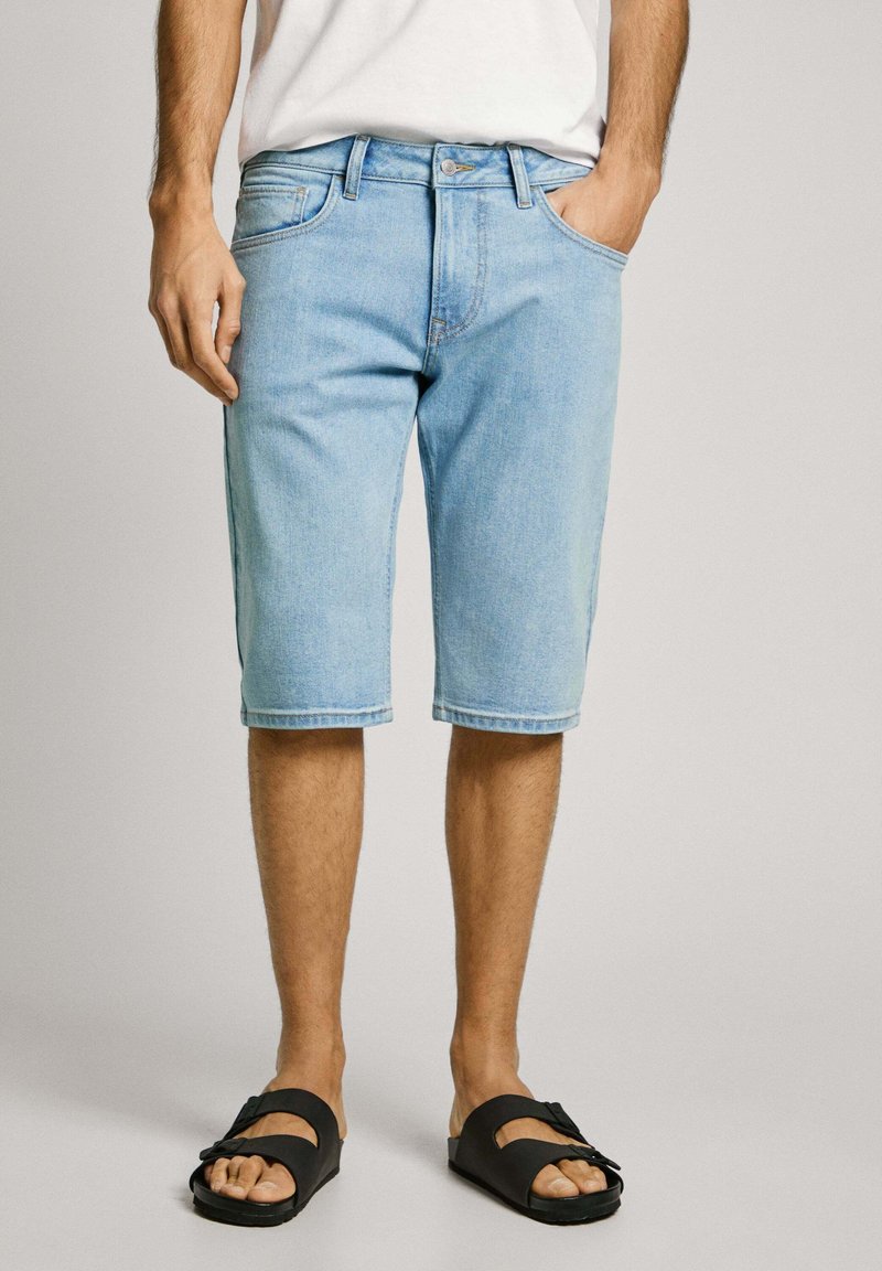 Pantalones cortos de mezclilla de color azul claro con corte recto, que cuentan con bolsillos laterales y un diseño clásico de cinco bolsillos, combinados con sandalias planas negras.