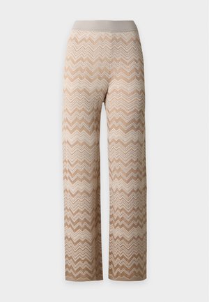 Frau steht und trägt eine schwarz-graue, zickzackmusterige, geknöpfte Strickjacke sowie beige, zickzackgemusterte, weite Hosen mit schwarzen Sandalen mit hohen Absätzen.
