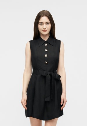 Young woman with straight brown hair wearing a sleeveless black dress with large square buttons and a tied waist belt, standing against a plain background.