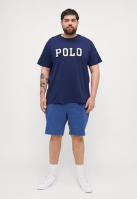 Tricou de culoare bleumarin cu inscripția "POLO" în litere albe îngroșate, asortat cu pantaloni scurți de culoare albastră deschisă. Modelul poartă adidași albi și șosete de lungime medie.