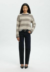 Jeune femme aux cheveux longs et bouclés, portant un pull rayé beige, un jean noir et des escarpins noirs à talons aiguilles, se tenant contre un arrière-plan uni.