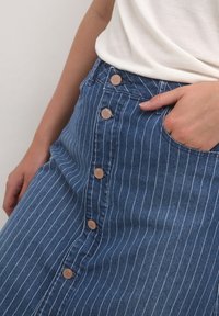 Jeanskjol med vertikala vita ränder, med knappar framtill, sidoficka och smal midja. Tyget är blått med en lätt struktur.