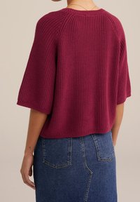 Svetr s krátkým rukávem ve tmavě burgundské barvě z pleteného materiálu; má žebrovanou strukturu a volný střih, kombinovaný s džínsovou sukní.