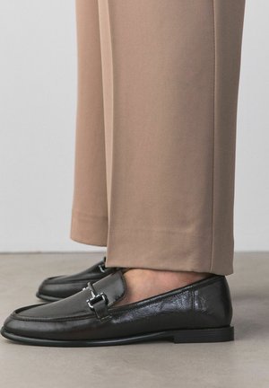 Mocassins en cuir noir avec détail en métal, portés avec un pantalon beige sur mesure sur un sol en béton.