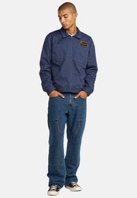 Veste bleu marine avec fermeture éclair, deux poches poitrine et un logo en patch. Assortie à un jean en denim bleu ample et des baskets noires.