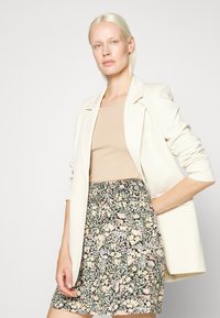 Blazer creme com lapelas ajustadas sobre uma blusa bege, combinado com uma saia com padrão floral em cores escuras com detalhes em rosa e amarelo.