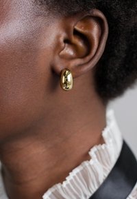 Guld, ovalformet hoopsmykke vist på en persons øre, der reflekterer lys med en glat, skinnende overflade mod mørk hud.